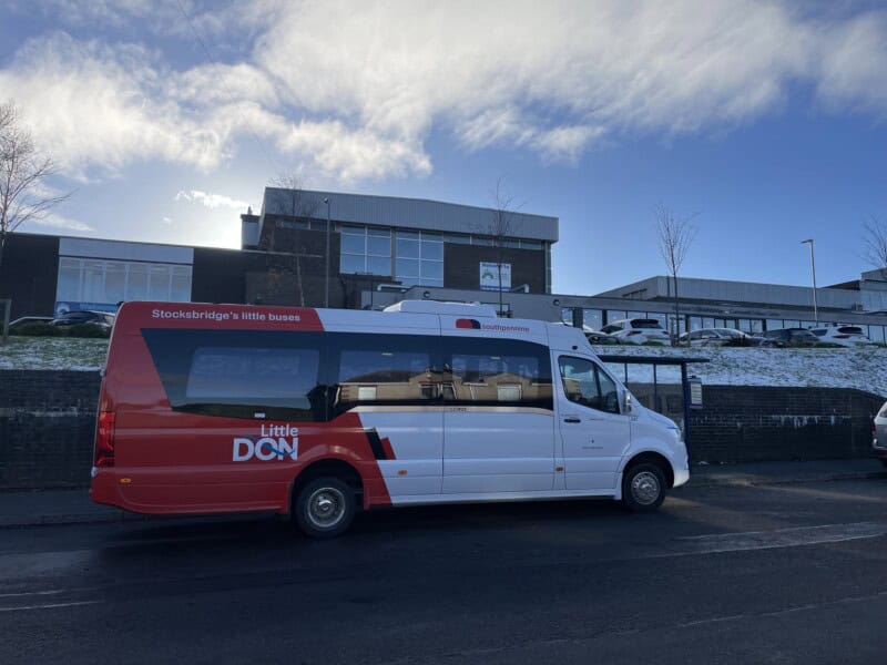 A red and white Little Don bus parked at a bus stop outside Stocksbridge Leisure Centre with a blue sky and frost covered grass in the background.